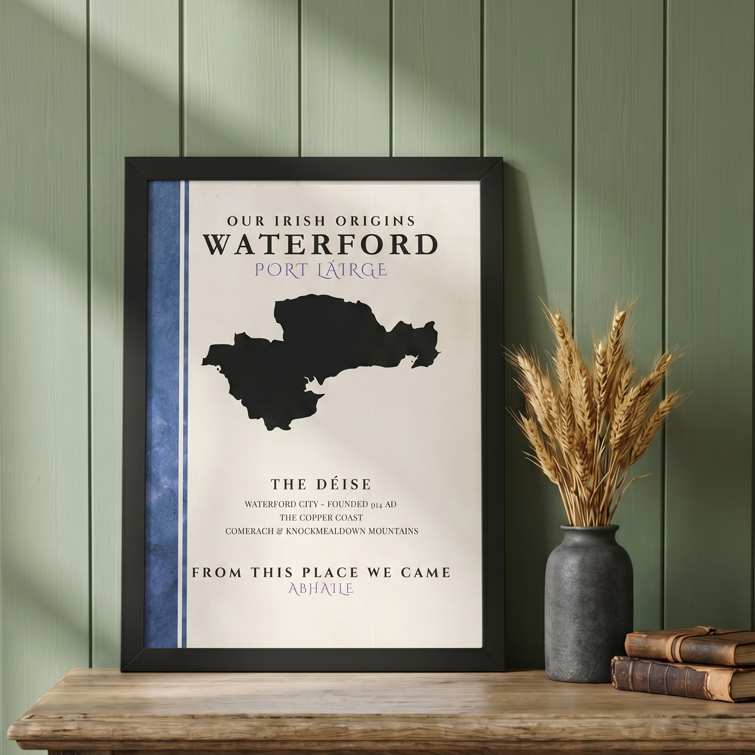 A framed typographic art print leaning on a wooden table against a green paneled wall, next to a gray vase with dried wheat and stacked books. The print reads 'Our Irish Origins Waterford Port Large' with a black map silhouette and blue side panel.