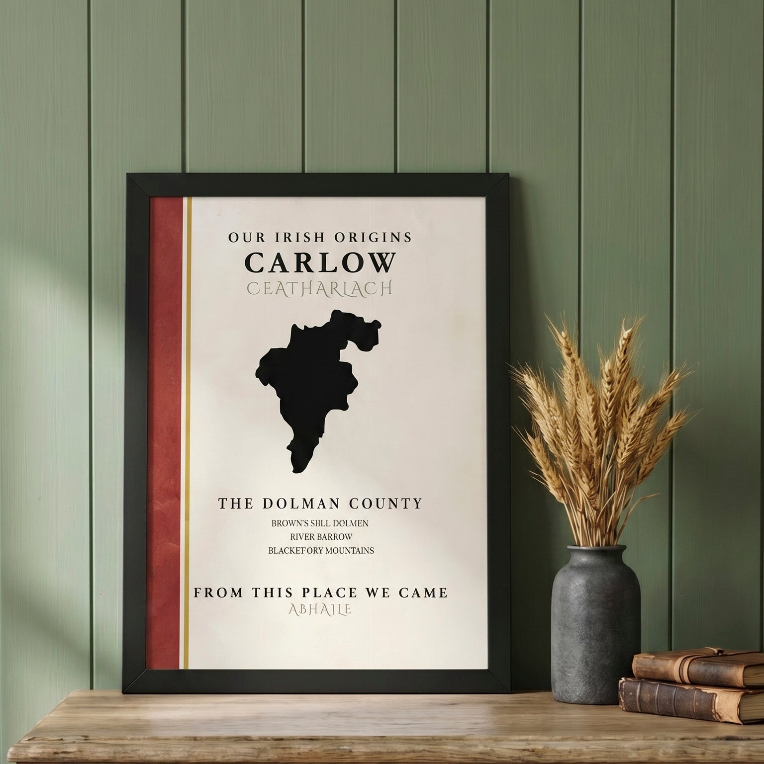 A framed personalized Irish origin poster for Carlow, displayed on a wooden table next to a vase of dried wheat and books, set against a green wood-paneled wall.